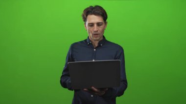 Laptop tutan ve ekrana bakan genç adam, tepki verirken yeşil stüdyoda alnına dokunur; sürpriz endişe.