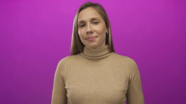 Canlı pembe bir arka plana gülümseyen, kahverengi bir kazak giyen, sakin ve kendinden emin bir ifade sergileyen bir kadın..