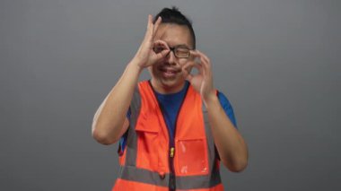 Yansıtıcı turuncu yelek giyen bir adam stüdyo gri arka planında el dürbünü hareketi yapıyor..