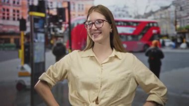 Gözlüklü bir kadın ve elleri kalçasında bağlı bir gömlek kalabalık bir caddede dil uzatıyor ve arka planda kırmızı bir otobüs var..