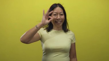 Soluk sarı tişörtlü kadın sağ eliyle ok işareti gösteriyor ve parlak sarı stüdyo duvarına gülümsüyor; kendine güven iyimserliği.