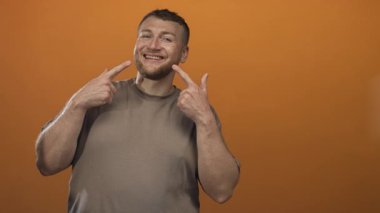 Genç adam parmaklarını dişlerini gösteriyor ve turuncu stüdyoda gülümsüyor, sakallı ve sıradan; mutluluk güveni.