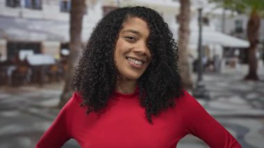 Kırmızı elbiseli kadın, açık havada, güneş ışığı altındaki şehir caddesinde elleri kalçalarında gülümserek poz veriyor..