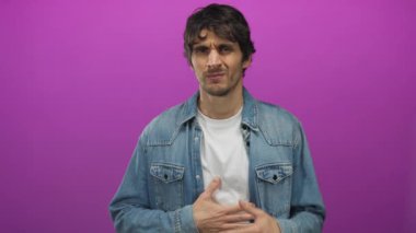 Mor bir stüdyo ortamında elleri çenesinde kot ceketli genç adam, dalgın bir poz alıyor; tefekkür.