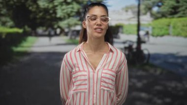 Kolları arkada olan kadın yeşil parkta dikilirken gözlük ve çizgili gömlek takıyor; şüphecilik şüphecilik şüpheciliğe şüphecilik şüpheciliğe şüpheye düşürür.