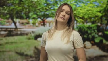 Parktaki sarışın kadın gülümserken ve ciddileşirken yeşil ağaçlarla dolu bir ortamda çeşitli duyguları yakalıyor..