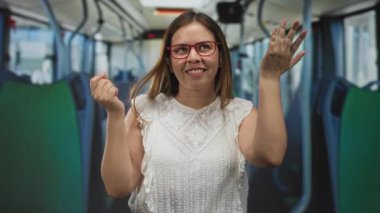 Kırmızı gözlüklü ve beyaz dantelli üst omuz silkme omuzlu bir kadın sokakta bir otobüsün içinde avuç içiyle gülümsüyor; neşeli bir merak.