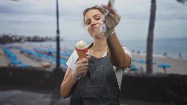 Plajda eldivenli dondurma külahını tutan kadın parmak kalp hareketi yapıyor; neşeli yaz neşesi.
