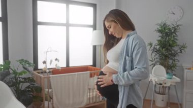 Bebek odasında karnını tutan genç sarışın hamile bir kadın. Etrafı beşik ve saatle çevrili. Huzurlu bir ev ortamı yaratıyor.