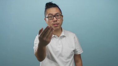 Beyaz polo giyen ve gözlüklü bir adam soluk mavi stüdyoda sağ elini kameraya doğru çekiyor; dostça bir davet..