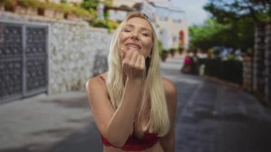 Kırmızı dantelli sutyen giyen sarışın kadın gülümsüyor ve güneş ışığında parlayan kaldırım taşı caddesinde öpücük yolluyor..