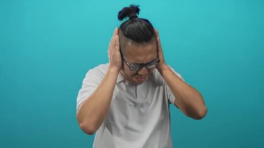 Elleriyle kulaklarını kapatan ve turkuaz bir stüdyoda somurtan adam beyaz polo ve gözlük takıyor, saçları topuz düğümüne bağlanmış, rahatsızlık duyuyor..