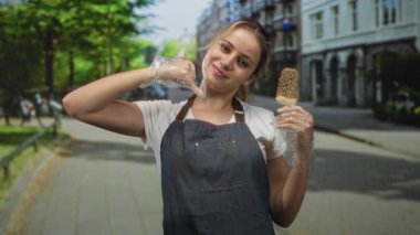 Dondurma tutan ve sokakta telefon jesti yapan bir kadın, önlük ve plastik eldiven takıyor; kendine güvenen dostane servis.