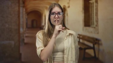 Taş bina koridorunda sessiz kalmak için parmaklar dudaklara genç sarışın kadın; sessiz içsel gizem.