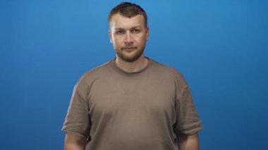Sakallı, çıplak kollu, mavi stüdyoda kameraya bakan, kahverengi tişörtlü, sakin bir adam..