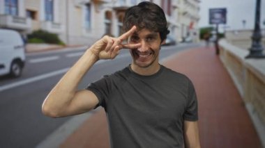 Güneşli bir şehir caddesinde göz ucuyla barış işareti yapan adam binalarla dolu, iyimserlik dolu..