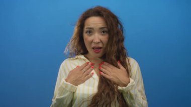 Mavi stüdyoda elleri göğsünde ve ağzı açık bir kadın çizgili gömlek ve kırmızı tırnak cilası giyiyor..