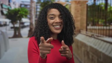 Kırmızı tişört giyen Afro-Amerikalı kadın şehir merkezindeki caddede kameraya işaret ediyor..