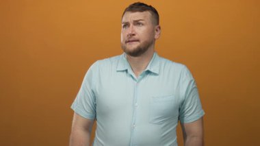 Kaşlarını kaldırmış ve şaşkın bakışlı bir adam açık mavi gömlek giyiyor ve turuncu stüdyo ortamında kısa kollu; belirsizlik yansıması.