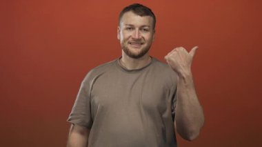 Turuncu stüdyo arka planında başparmağıyla işaret eden adam, görünür sakal ve günlük tişörtle gülümsüyor; dostça yaklaşılabilir ruh hali.