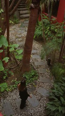 Lüks bir tatil köyü ortamında verimli bir tropikal bahçede yürüyen bir kadın, sakin atmosferi ve etrafındaki doğal güzellikleri palmiye ağaçları ve çakıllı yollarla vurguluyor..