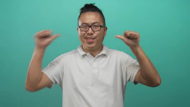 Beyaz polo giyen bir adam iki başparmağını da göğsüne doğru uzatıyor ve elleri tepe rengi stüdyoda havaya kalkıyor..