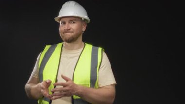 Beyaz kasketli ve neon renkli yelek giyen adam, stüdyo siyah arka planda elleri göğsüne kenetlenmiş; güven planlaması.