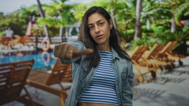 Tatil yerindeki kameraya doğru işaret eden, çizgili gömlek ve kot ceket giyen, kendine güvenen bir kadın..