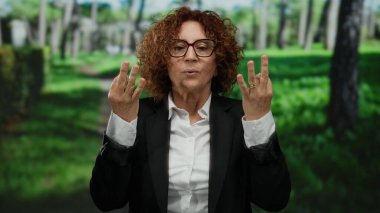 Parkta nefes egzersizi yapan İspanyol kadın gözlük ve takım elbise takıyor, açık havada yemyeşil ağaçlar ve çimlerle çevrili..