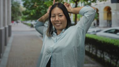 Görünüşte bulanık ağaçlar ve arabalarla bir şehir caddesinde gülümseyen ve başının arkasına barış işareti yapan bir kadın..