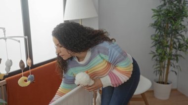 Genç bir kadın rahat yatak odasında renkli süveter ve kıvırcık saçlarıyla bebek karyolasını ayarlıyor.