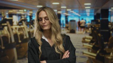 Spor salonunda kollarını kavuşturmuş bir kadın sinirli görünürken, üniforma giyen genç sarışın yargıç içeride spor salonu duvarının yanında duruyor..