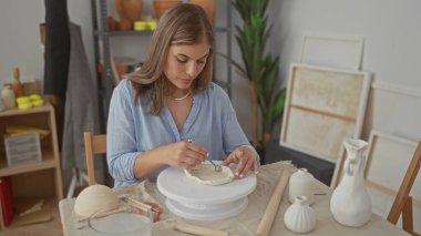 Sarışın kadın kil kaseyi şekillendirmek için çıplak el ve metal heykel aleti kullanıyor..