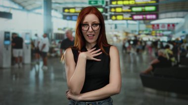 Havaalanında gülümseyen kızıl saçlı bir kadın bulanık yolcu ve uçuş tabelalarıyla dolambaçlı terminalde poz veriyor. İçerideki hareketli seyahat atmosferini yakalıyor..
