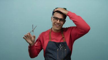 Gözlük ve önlük takan genç adam, kuaför ya da kuaför olarak yeteneklerini gösteren izole edilmiş mavi duvar arkaplanına karşı makas ve makas tutuyor.