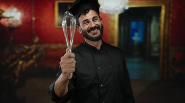 Şef gibi giyinmiş İspanyol sakallı bir adam sanat galerisinde çırpıcıyla kırmızı duvarlar ve dekoratif çerçevelerle müze ortamında aşçılık hevesi sergiliyor..