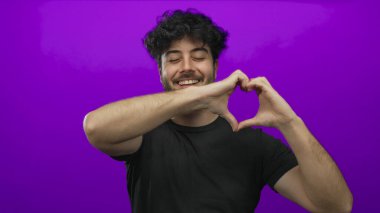 Sakallı genç İspanyol adam kalp hareketi yapıyor. Mor arka plana gülümsüyor. Sevgi ve pozitifliği ifade ediyor.