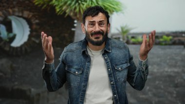 Kot ceketli İspanyol sakallı adam yeşil ağaçlar ve rahat şehir manzarasıyla çevrili bir parkta omuz silkiyor..