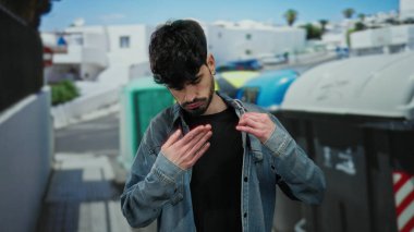 Siyah saçlı, kot ceketli, yakalı, beyaz binaların ve renkli kutuların bulanık arka planıyla şehir sokağında duran genç adam..