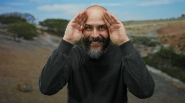 Sakallı ve dövmeli bir adam doğa ile etkileşim ve oyuncu ifadeler sergileyen manzaralı bir parkta jestler yapıyor..