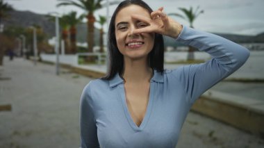 Genç İspanyol kadın deniz kenarında bulutlu gökyüzünün altında palmiye ağaçlarıyla zafer jesti yapıyor. Mavi kıyafetiyle neşe ve pozitifliğini ifade ediyor..