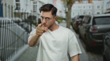 Sakallı genç bir adam şehir caddesinde kameraya doğru hareket ediyor, günlük beyaz tişört giyiyor, şehir dışında yaşıyor, dinamik bir duruşla ciddi duygular ifade ediyor..