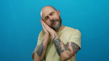 Sakallı ve dövmeli bir adam, izole edilmiş mavi arka plana karşı gözleri kapalı, sakin ve huzurlu bir hava yayıyor..