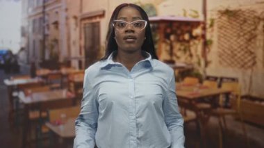 Gözlüklü ve mavi gömlekli kadın, sokak kafesinde masa ve sandalyeli el kaldırıyor..