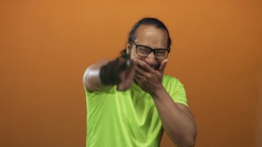 Gözlüklü bir adam stüdyoda turuncu arka plana gülerken eliyle ağzını kapatıyor..