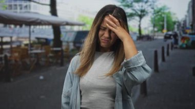 Elleri alnında bir kadın sokakta restoran terasının yanında, gün ışığında duruyor ve yüzünü ekşitiyor; rahatsız edici baş ağrısı.