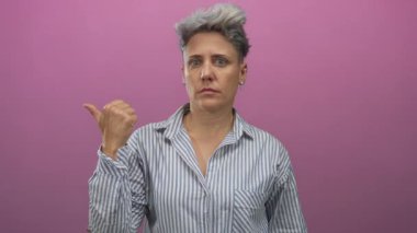 Kısa gri saçlı, çizgili tişörtlü beyaz bir kadın stüdyoda sessizlik için parmaklarını dudaklarına kaldırıyor..