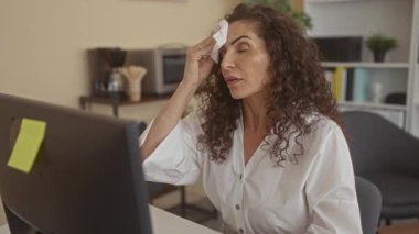 Genç İspanyol kadın, modern ofis binasındaki bilgisayar masasında çalışırken alnını siliyor..