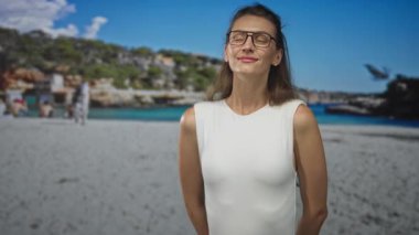 Gözleri kapalı gülümseyen bir kadın, stüdyo plajında beyaz kolsuz bir elbise içinde görünen çıplak omuzlar; dinginlik..