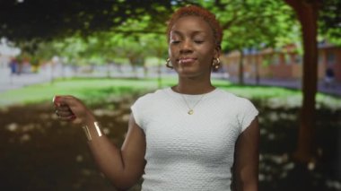 Beyaz bluz ve altın halkalı küpeler takan kadın, güneşli yeşil bir parkta kırmızı tırnaklı sıkılmış bir yumruğu kaldırıyor..